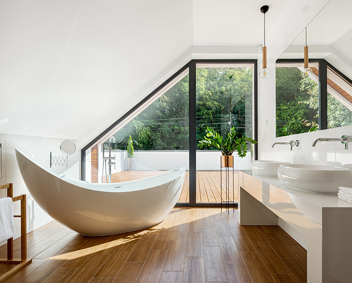 A modern bathroom featuring a stylish freestanding bathtub, wooden flooring, and large windows that offer a view of greenery outside, creating a bright and inviting space.